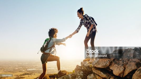 Woman helping her friend to climb the cliff and reach the top of mountain. Friends helping each other during hiking a mountain.